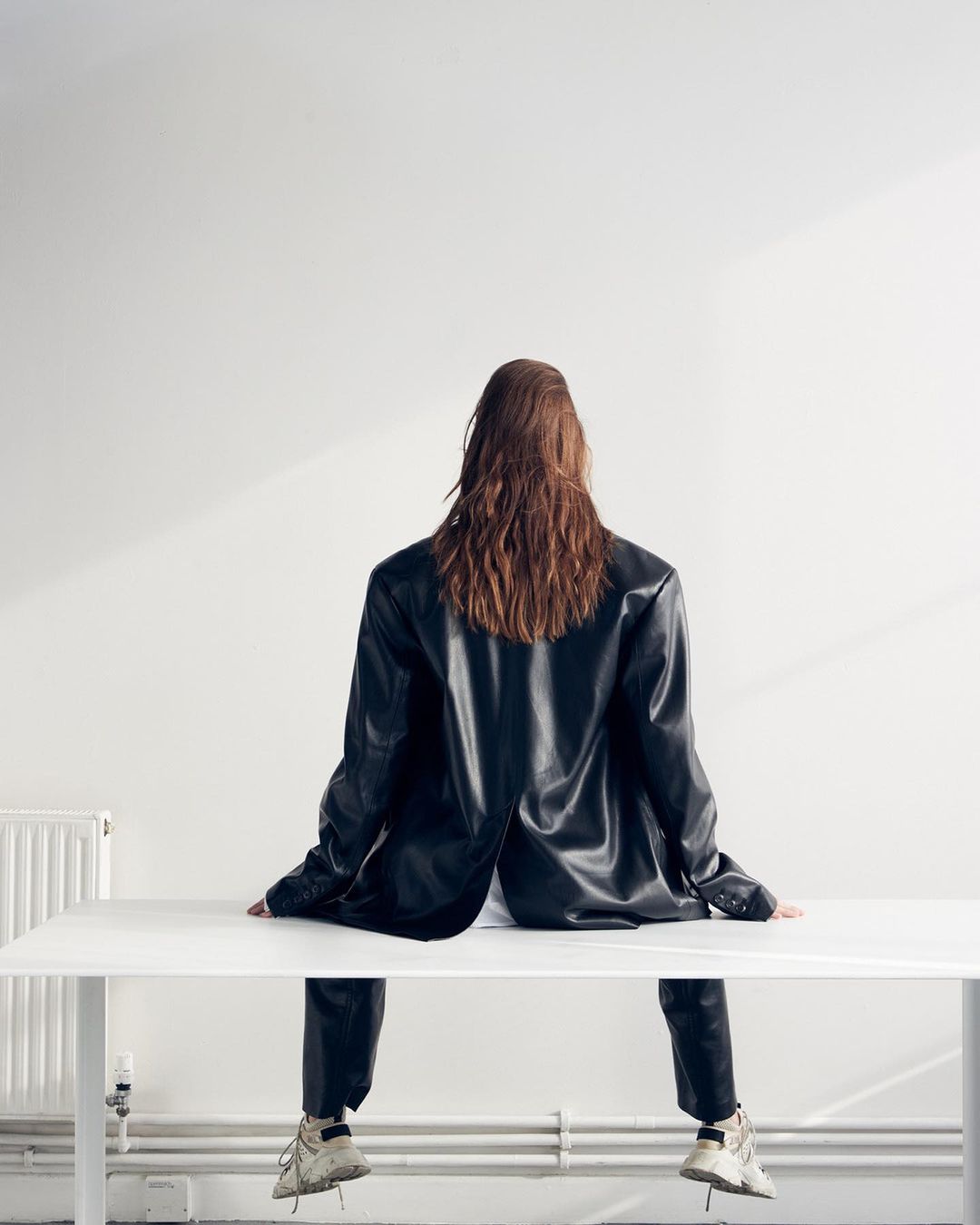 Model seen from behind wearing a black leather blazer and trousers seated on a white table against a plain wall