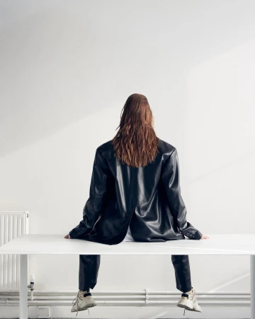 Model seen from behind wearing a black leather blazer and trousers seated on a white table against a plain wall