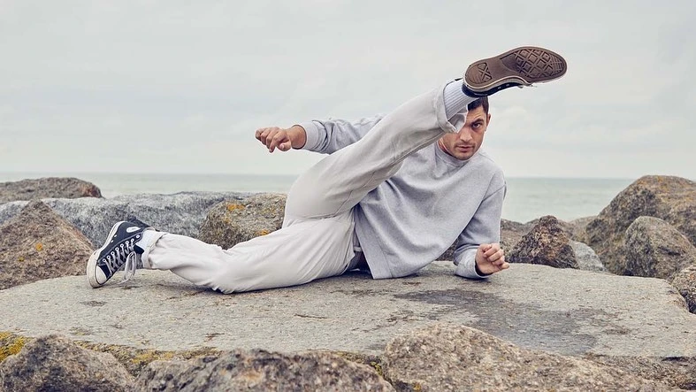 Athletic man in casual wear poses on rocks by the sea, with one leg raised high, showcasing balance and flexibility in a natural setting.