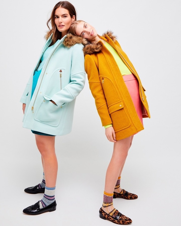 Two fashion models in pastel and mustard-colored winter coats with fur-trimmed hoods pose in a studio. They wear colorful socks and loafers.
