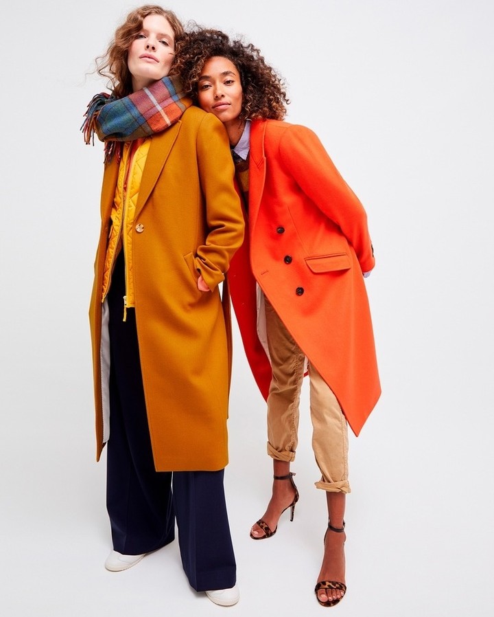 Two women pose in stylish fall coats against a bright background. One wears a mustard coat with a scarf, the other a double-breasted orange coat.