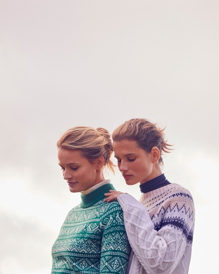 Two adult females wearing patterned, long-sleeved sweaters. One woman has her hand on the other's shoulder in a close-up outdoor shot.