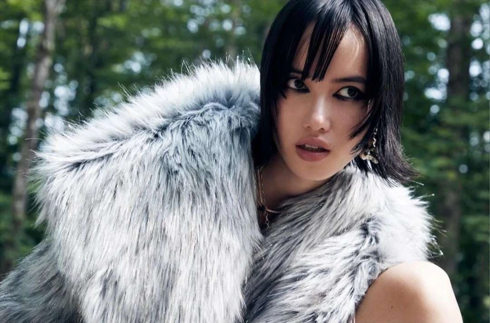 Portrait of an adult woman with bobbed hair wearing a faux fur coat, gazing off-camera in a forest environment with trees in the background.