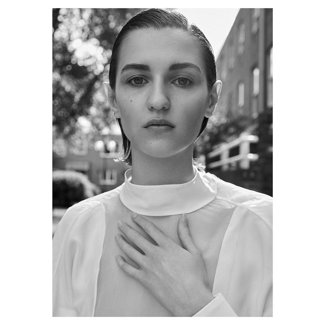 Monochrome portrait of a woman with slicked-back hair wearing a high-necked white top, posing outdoors with a building and trees in the background.