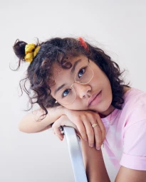 Close-up portrait of a young woman wearing wire-rimmed glasses and a pink shirt, leaning her chin on her crossed arms against a white background.