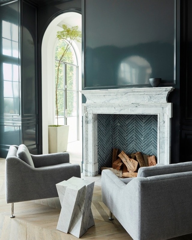 Living room interior with gray armchairs, geometric side table, a marble fireplace with wood, and high-gloss black paneling.