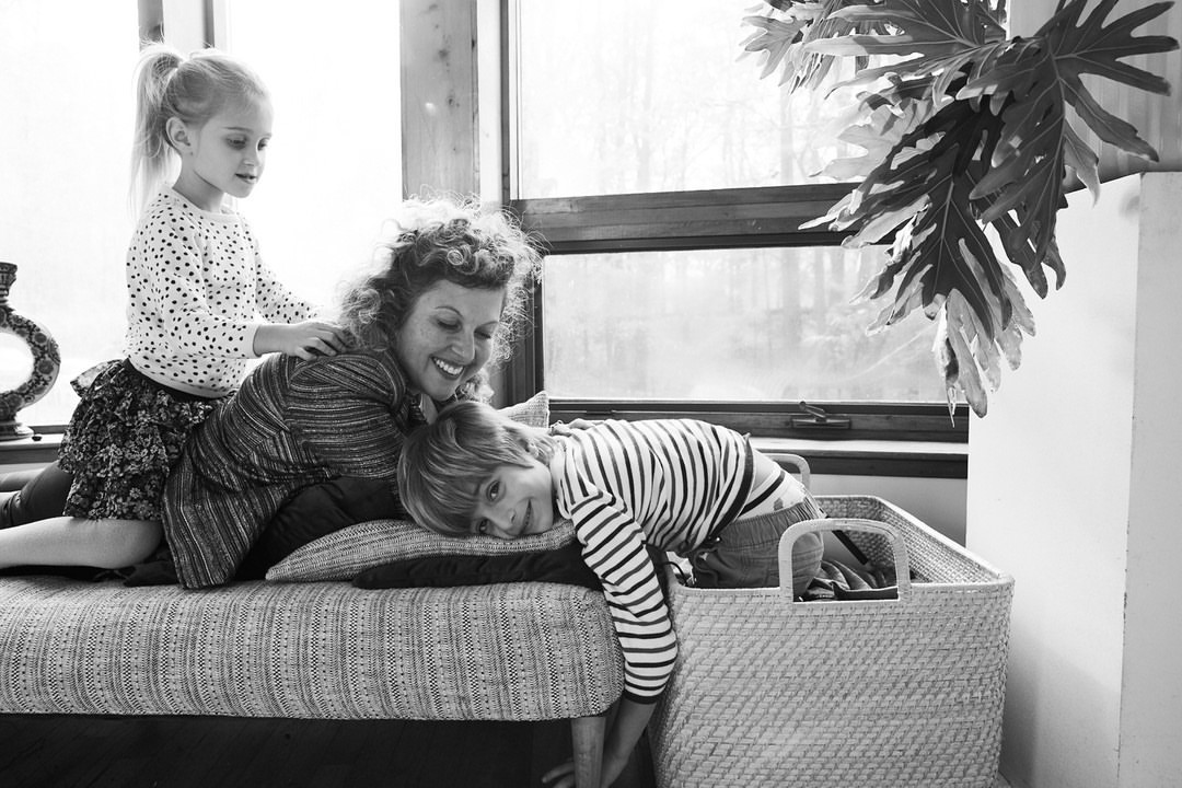 A mother and her two children are sitting on a cushioned bench near a window. The child is giving the mother a massage.