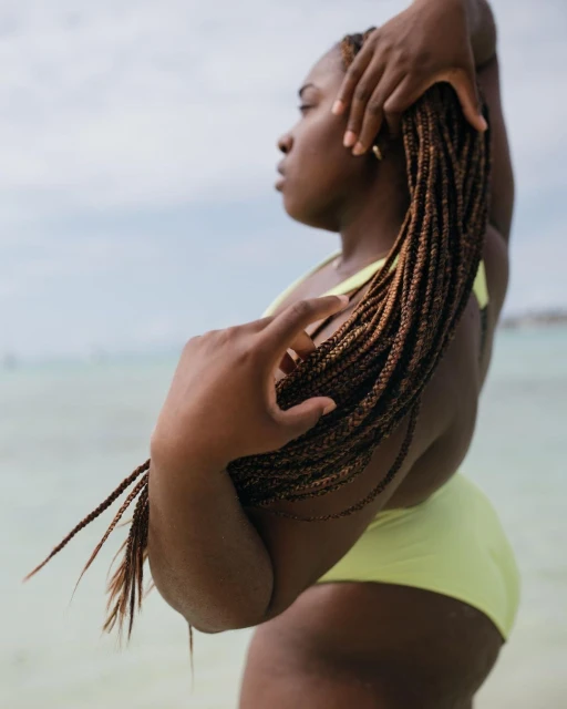 African American woman in a lime-green swimsuit, standing near the ocean with her hair in braids, posing in natural light.