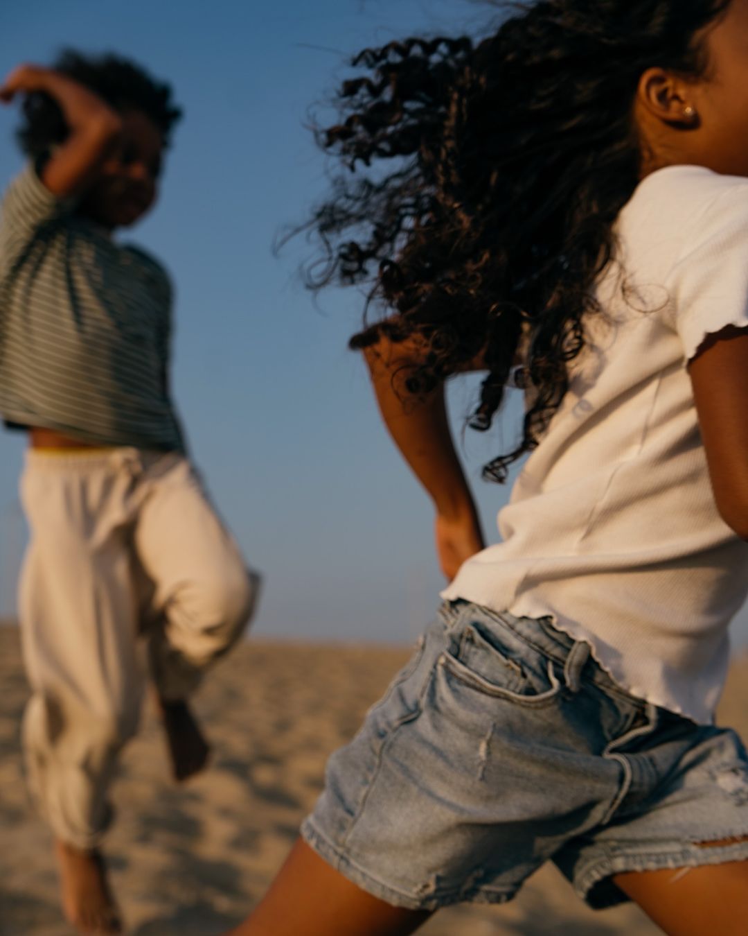 Two children, one wearing a white top and jean shorts, the other a striped top and beige pants, run on a sandy surface under a blue sky.