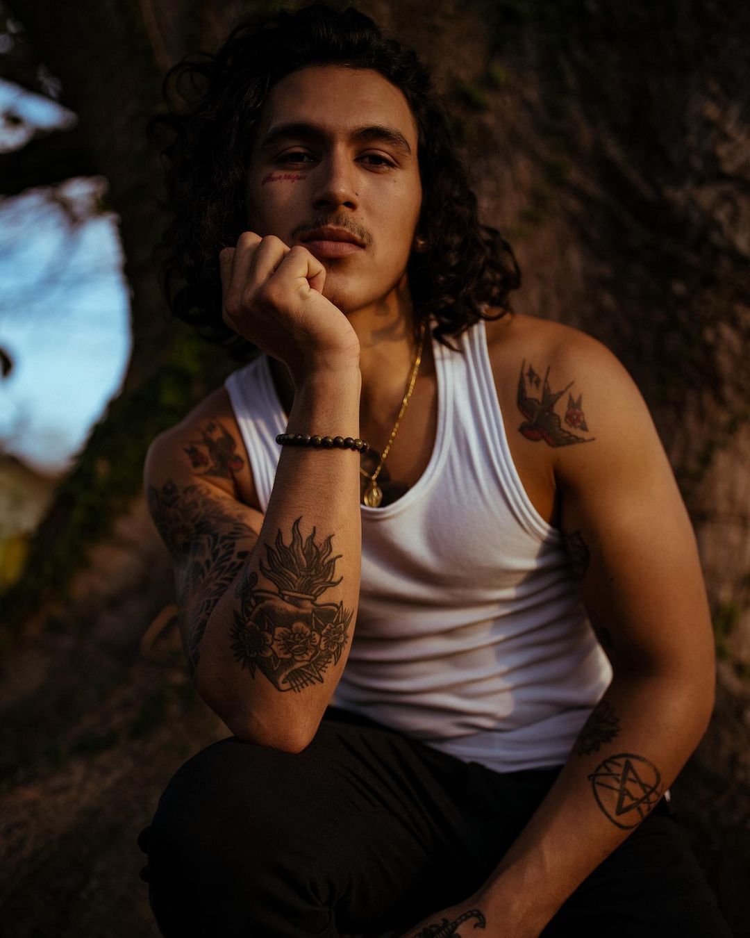 Portrait of a young adult man with tattoos wearing a white tank top, sitting outdoors with a tree trunk visible in the background.