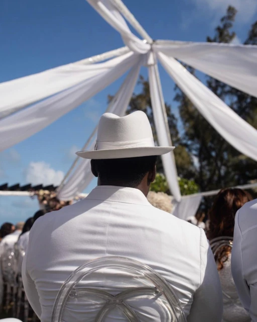 Back view of a person in white suit and hat at an outdoor event with white draping, under a bright blue sky with light cloud cover.