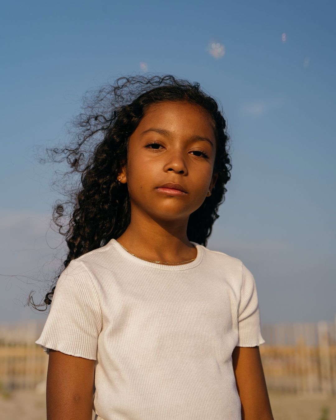 Portrait of a young girl with curly hair, wearing a white t-shirt, posing outdoors against a blue sky in natural light.