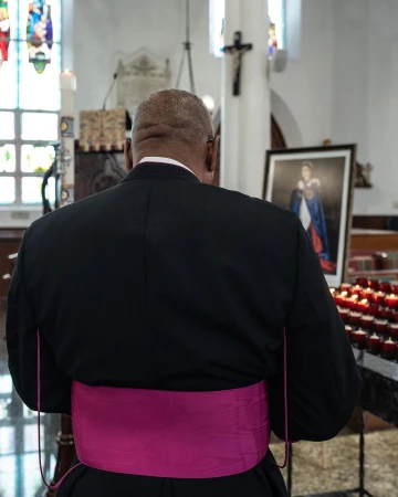 Clergy member in a black robe and pink sash stands inside a church with rows of lit candles and a portrait of a royal figure.