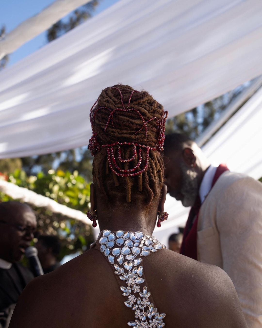 Woman in an elegant white dress with a jeweled neckline, facing away. A wedding party is visible in the soft-focused background.