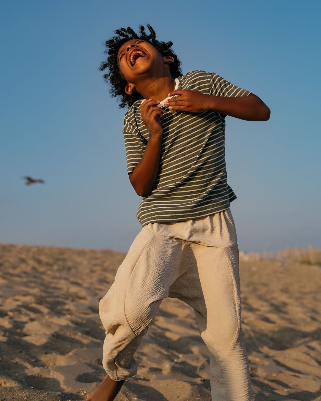 Young boy with curly hair laughs in a striped shirt on a sandy beach, under a clear blue sky, enjoying a bright, sunny day outdoors.