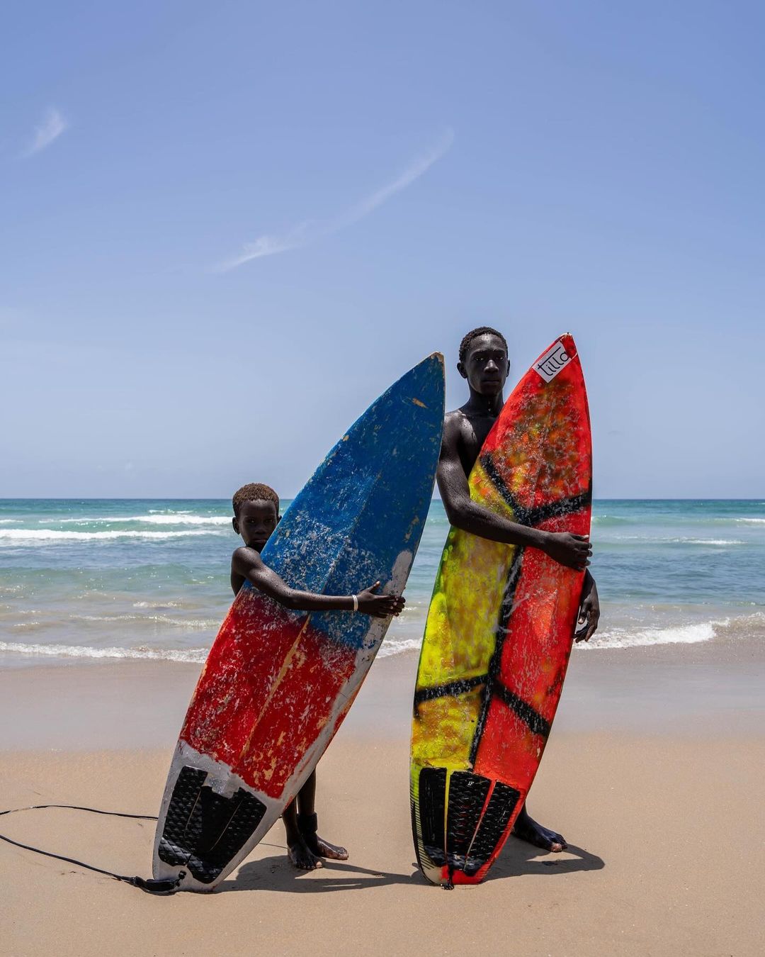 Two people on a beach holding colorful surfboards, one adult and one child, with ocean waves and clear sky in the background.