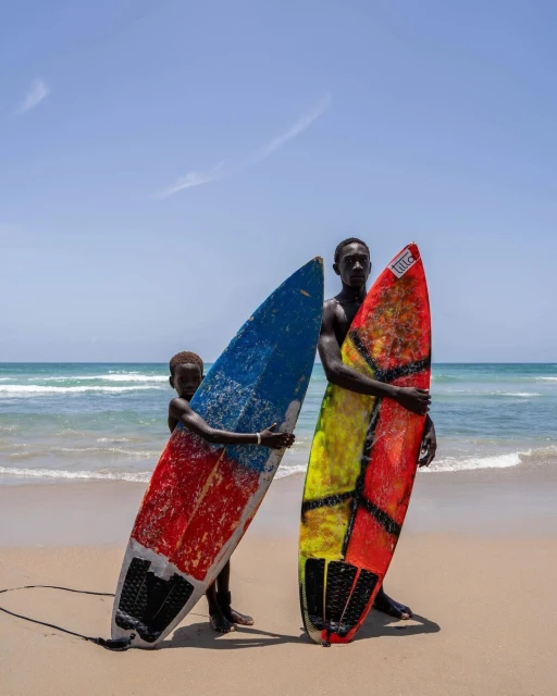 Two people on a beach holding colorful surfboards, one adult and one child, with ocean waves and clear sky in the background.