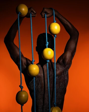 A muscular man with dark skin poses shirtless with yellow exercise balls suspended from blue ropes against an orange background.