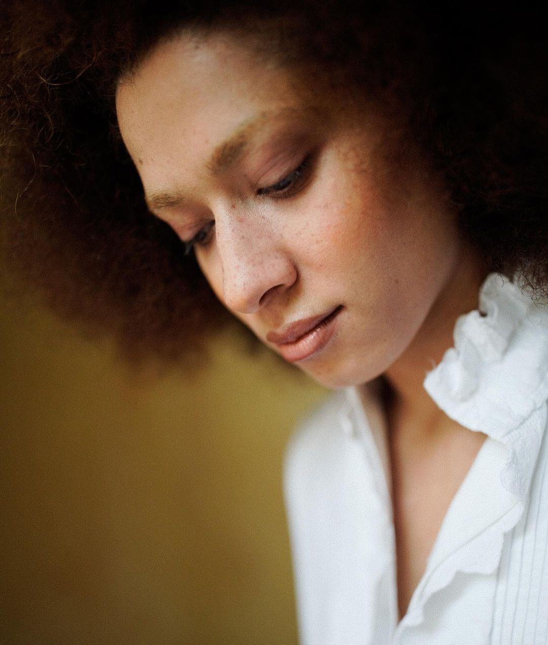 Close-up portrait of a young adult female with curly hair and freckles, wearing a white ruffled shirt, set against a neutral background.