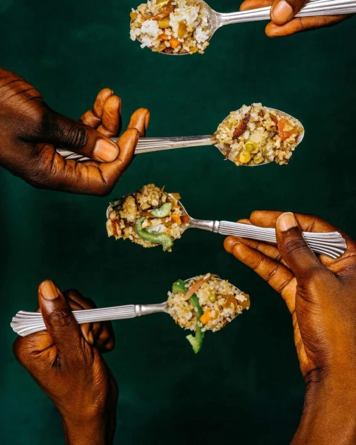 Dark-skinned hands holding silver spoons with colorful food against a dark green background in a studio food and beverage setting.