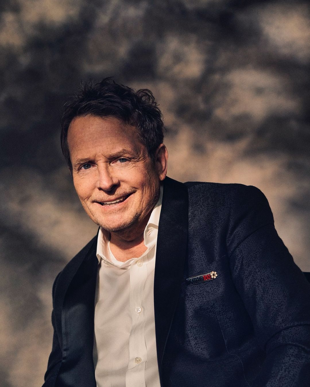 Portrait of Michael J. Fox wearing a black tuxedo jacket and white shirt. He smiles softly against a mottled gray background.