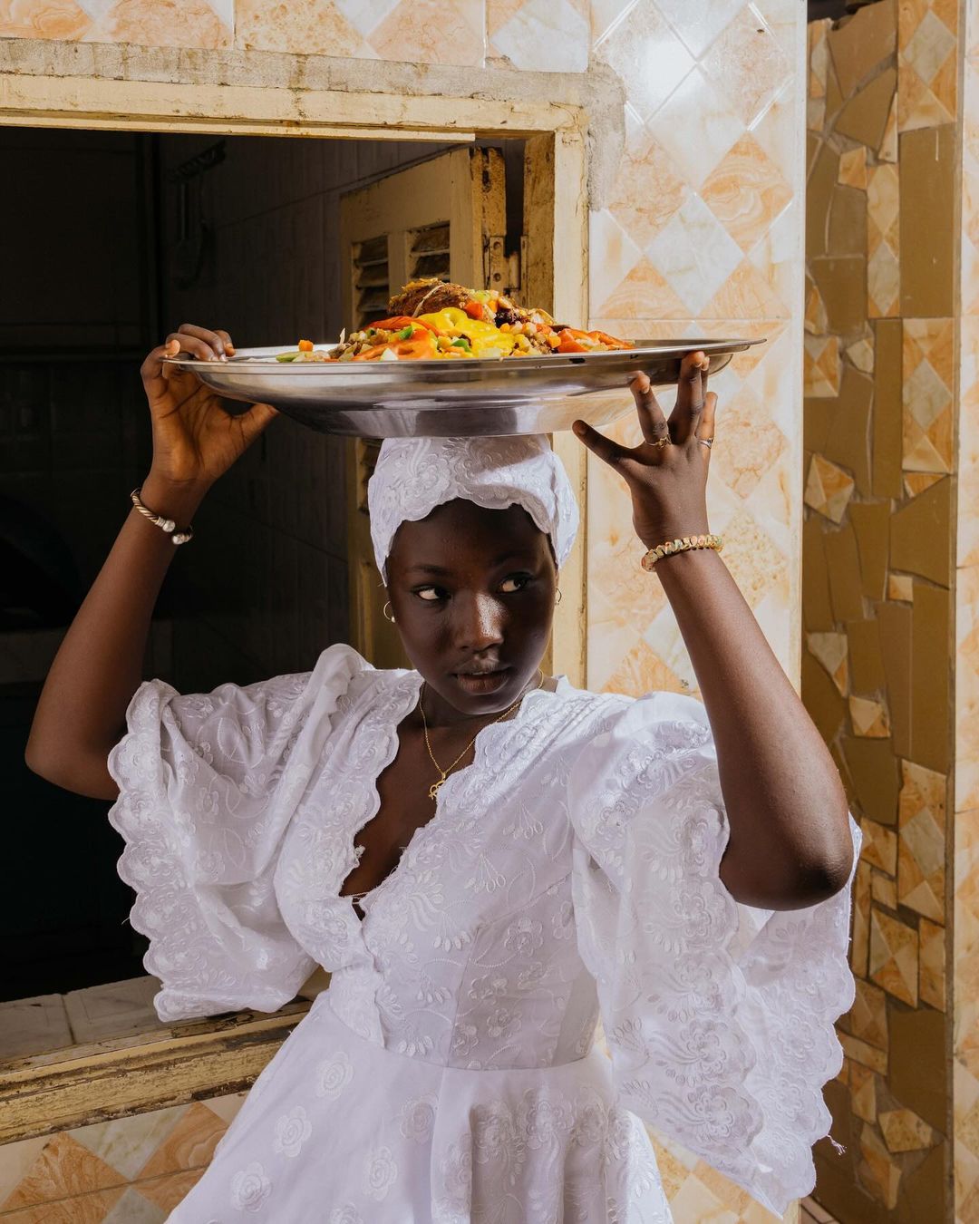 Dark-skinned woman wearing a white dress and head covering balances a tray of food on her head in front of a window with tile surround.