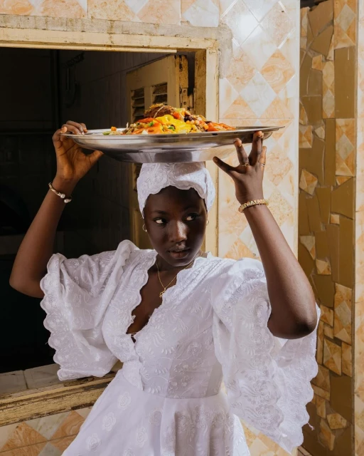 Dark-skinned woman wearing a white dress and head covering balances a tray of food on her head in front of a window with tile surround.