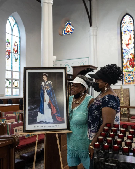 Two women in a church view a framed portrait of Queen Elizabeth II. Candles flicker in the foreground, stained glass windows are visible.