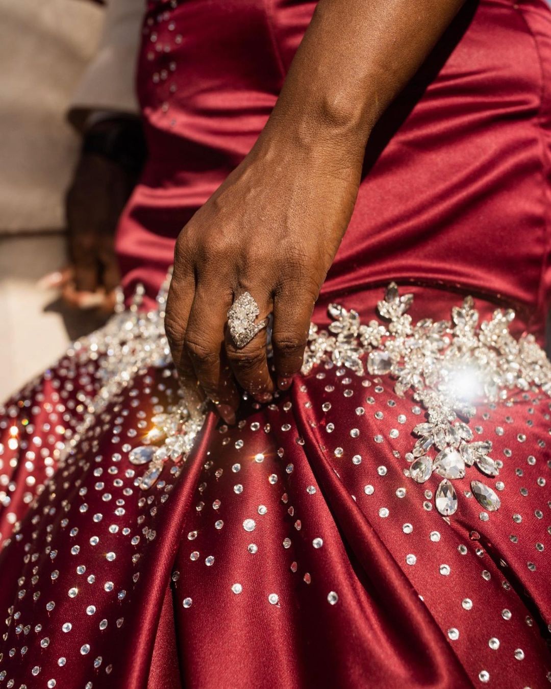 Close up of a dark-skinned woman's hand wearing a large jeweled ring resting on a wine-colored, rhinestone-studded gown with floral detailing.