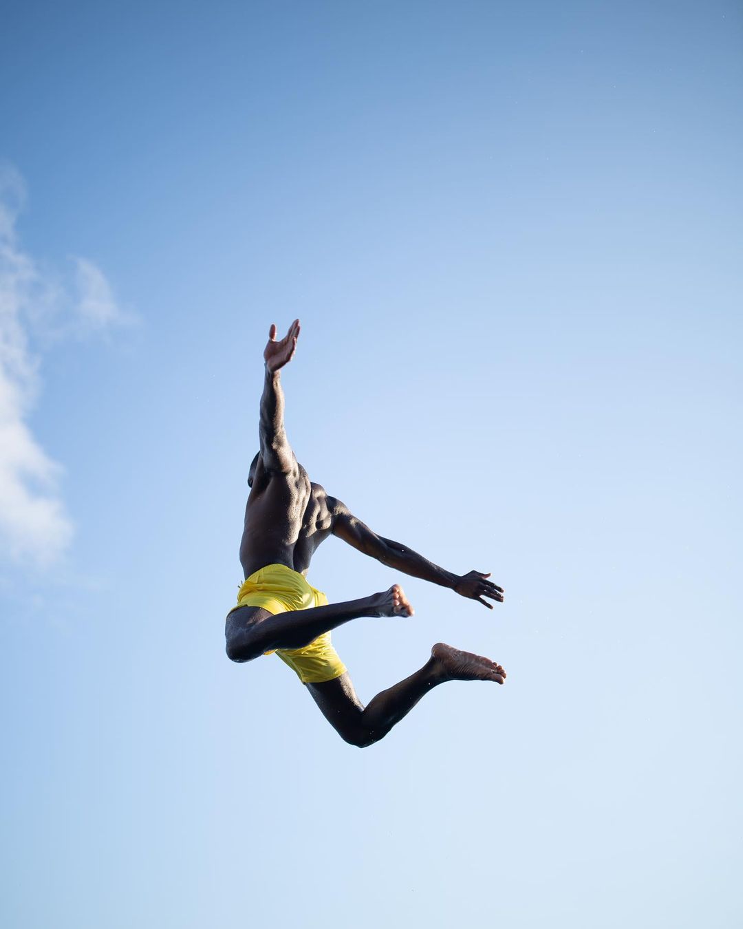 Athletic Black male jumping in the air with arms raised wearing yellow shorts against a clear blue sky with minimal white clouds.
