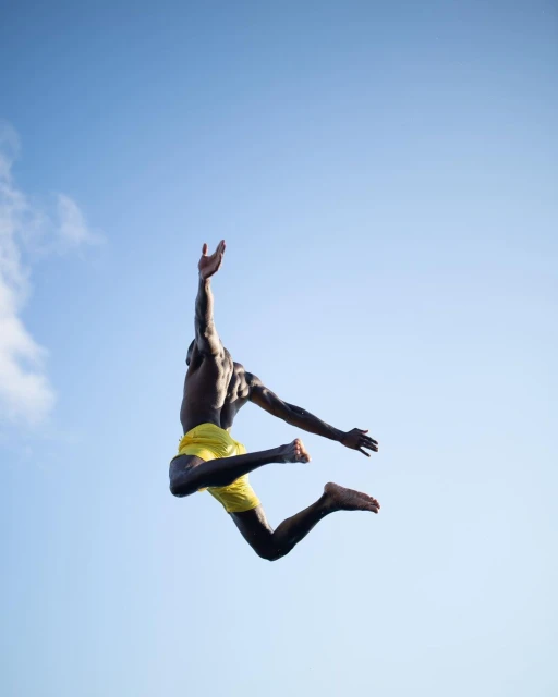 Athletic Black male jumping in the air with arms raised wearing yellow shorts against a clear blue sky with minimal white clouds.