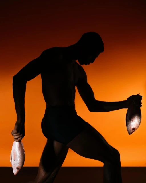 Silhouette of muscular man holding fish in each hand against an orange background in a studio with strong lighting.