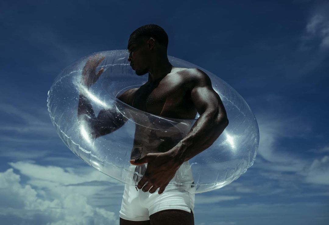 Athletic African American man posing with clear inflatable swim ring against a blue sky with sparse clouds on a sunny day. 