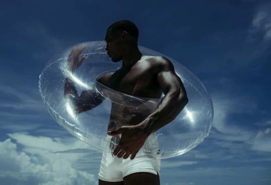 Athletic African American man posing with clear inflatable swim ring against a blue sky with sparse clouds on a sunny day. 