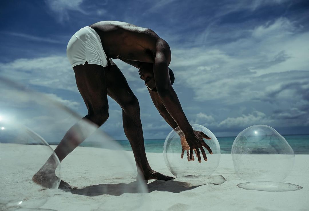An African-American adult male bends over to touch a clear plastic bubble on a sandy beach, with a blue sky and clouds visible above.