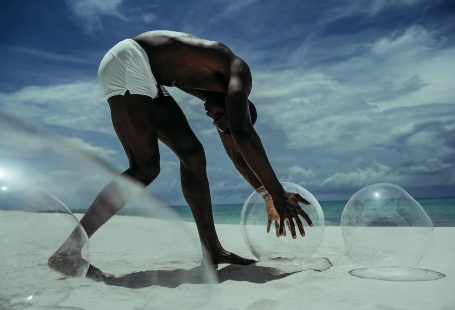 An African-American adult male bends over to touch a clear plastic bubble on a sandy beach, with a blue sky and clouds visible above.