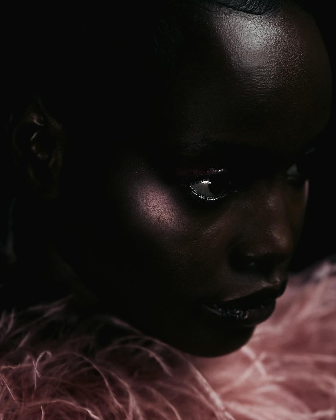 Close-up portrait of a woman with dark skin and glossy lips, wearing a pink textured garment, against a dark, shadowy background.