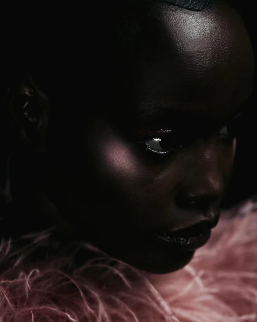 Close-up portrait of a woman with dark skin and glossy lips, wearing a pink textured garment, against a dark, shadowy background.