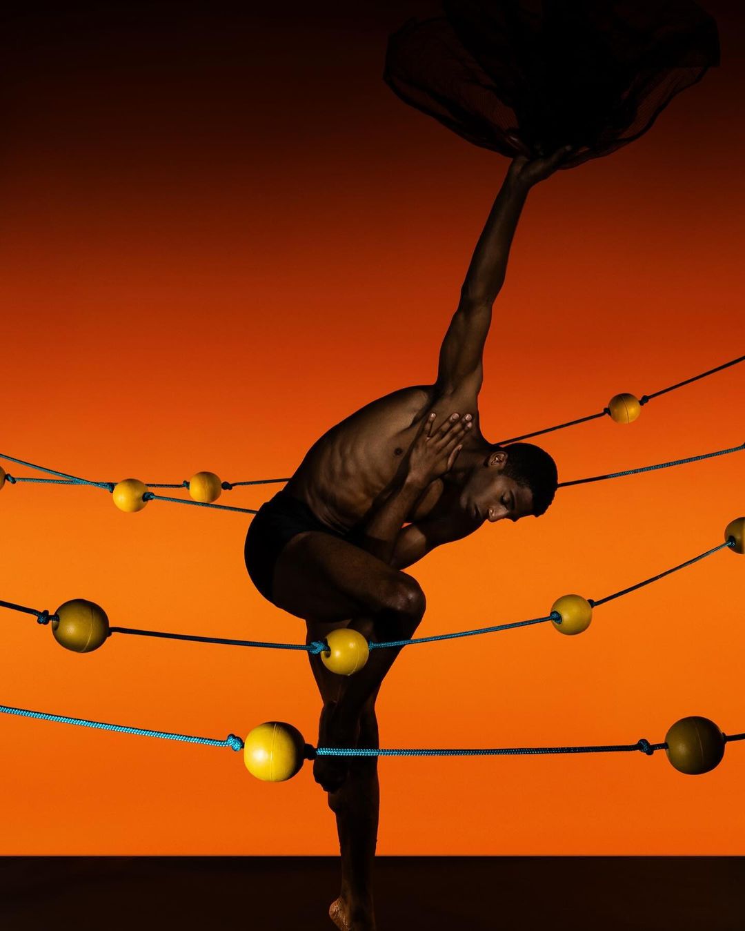 A dark-skinned athletic man balances in a challenging yoga pose against a warm orange backdrop, surrounded by ropes and yellow spheres.