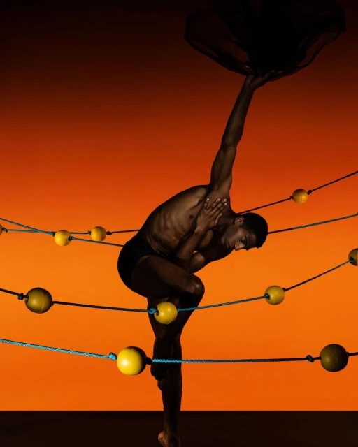 A dark-skinned athletic man balances in a challenging yoga pose against a warm orange backdrop, surrounded by ropes and yellow spheres.