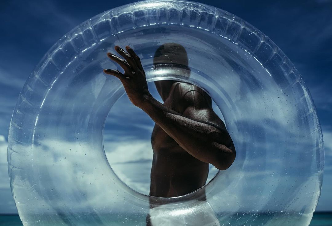 Adult male with dark skin stands on a beach, looking to the side through the center of a clear plastic inflatable swim ring.