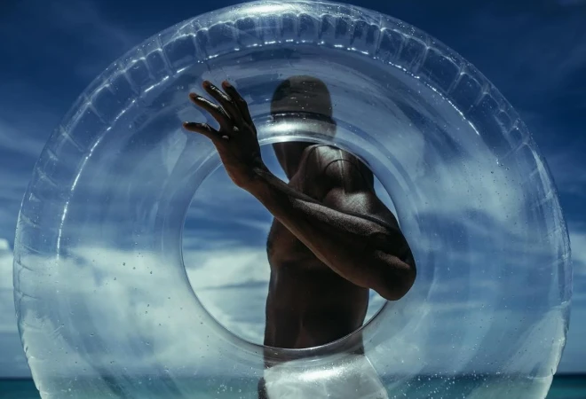 Adult male with dark skin stands on a beach, looking to the side through the center of a clear plastic inflatable swim ring.