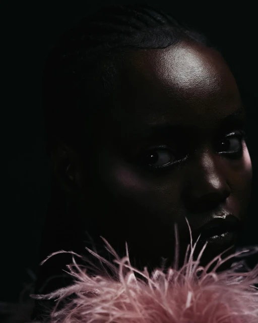 Close-up portrait of a Black woman with dark hair and glossy lips, gazing sideways against a dark background and wearing a feathered collar.