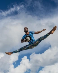 Black man mid-air in a dramatic split jump against a bright blue sky filled with white clouds, holding a hat.