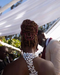 Rear view of a Black woman with locs and red beaded hair accessories wearing a crystal-embellished halter dress.