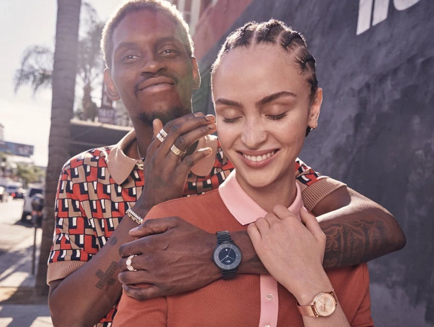 Smiling couple outdoors in sunlight.  An African-American man embraces a Caucasian woman. Watches on their wrists. Geometric shirt, blue wall.