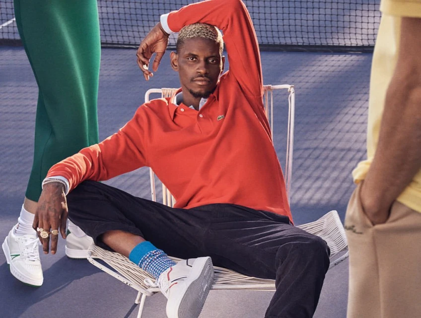 A blonde man in a red shirt poses on a white chair, with others nearby on a blue court surface, creating a fashionable, sporty editorial scene.