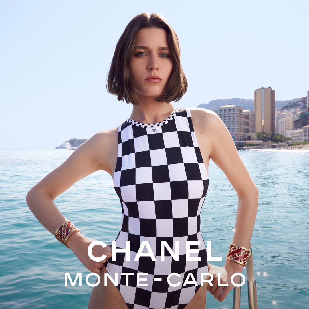 Woman poses in a black-and-white checkered one-piece swimsuit with Monte Carlo buildings and blue water background on a sunny day.