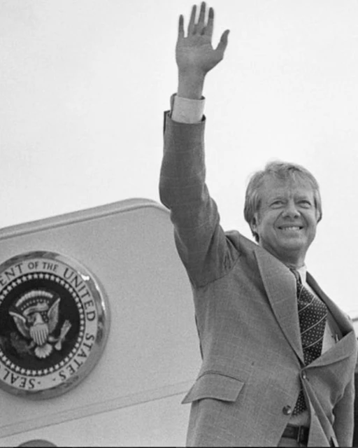 Monochrome portrait of Jimmy Carter waving with a smile, wearing a suit and tie with an airplane featuring the presidential seal visible.