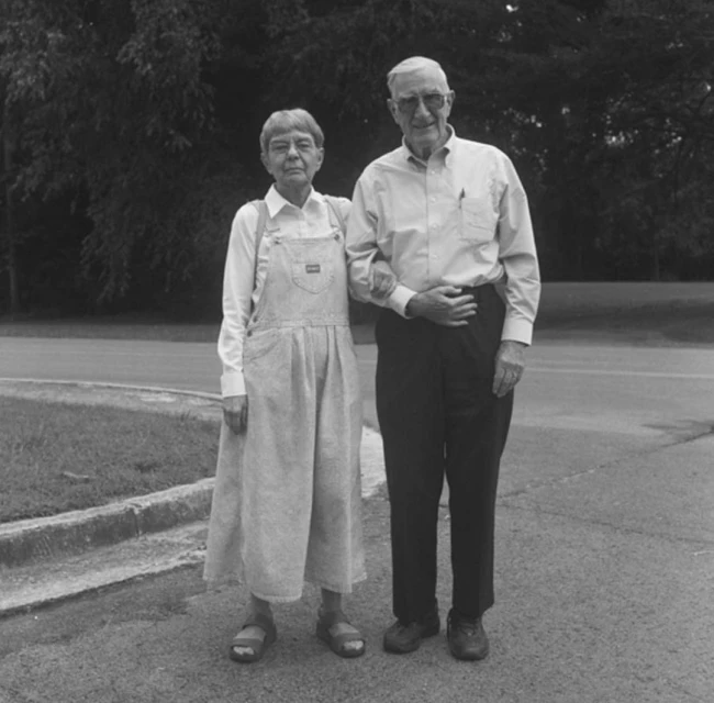 Elderly couple posing outdoors on a sunny day, standing on a road near a grassy area and trees; the woman wears overalls and sandals.